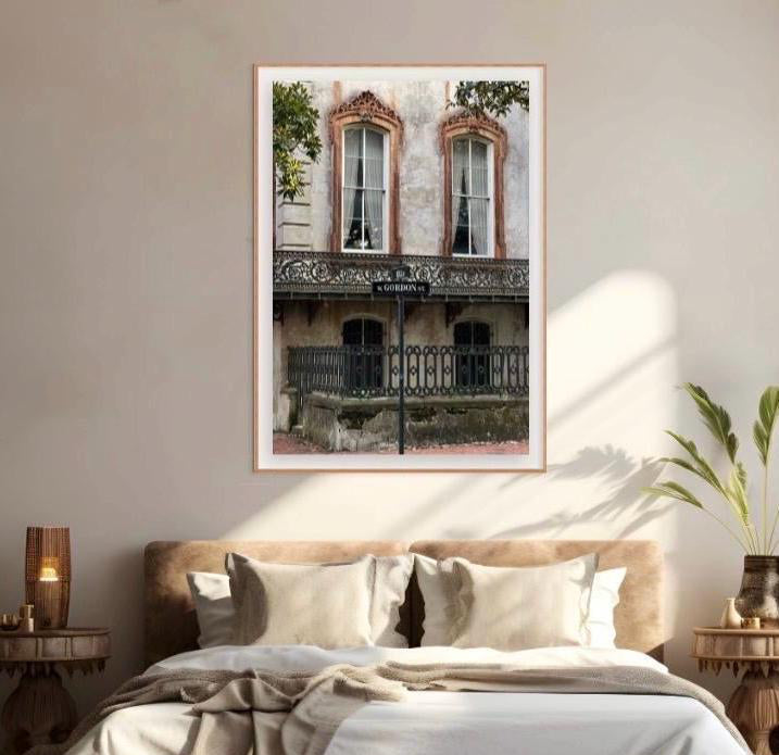 A framed wall art print depicting a vintage Parisian-style building known as the Noble Hardee Mansion in historic Savannah, Georgia, on Gordon Street facade with arched windows and a black wrought-iron balcony, hung above a neatly made bed in a softly lit bedroom.