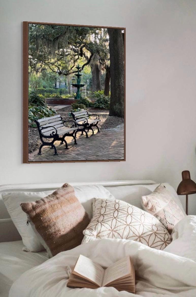 A framed wall art print depicting a park scene in Lafayette Square in historic downtown Savannah, Georgia, with benches, trees, and a fountain, displayed above a bed with white bedding and decorative pillows in a bedroom setting.