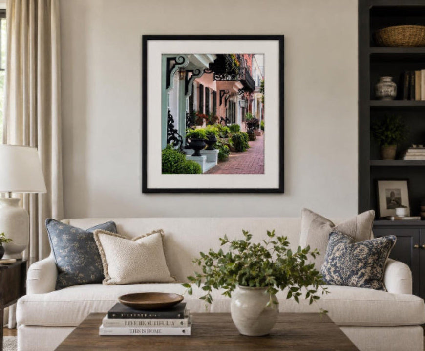 A framed art print depicting a charming European street with classic buildings and greenery, styled on a beige wall above a white sofa in a modern living room setting. Photo captured in historic Savannah, Georgia, and shows rainbow row-style houses similar to Charleston, South Carolina.